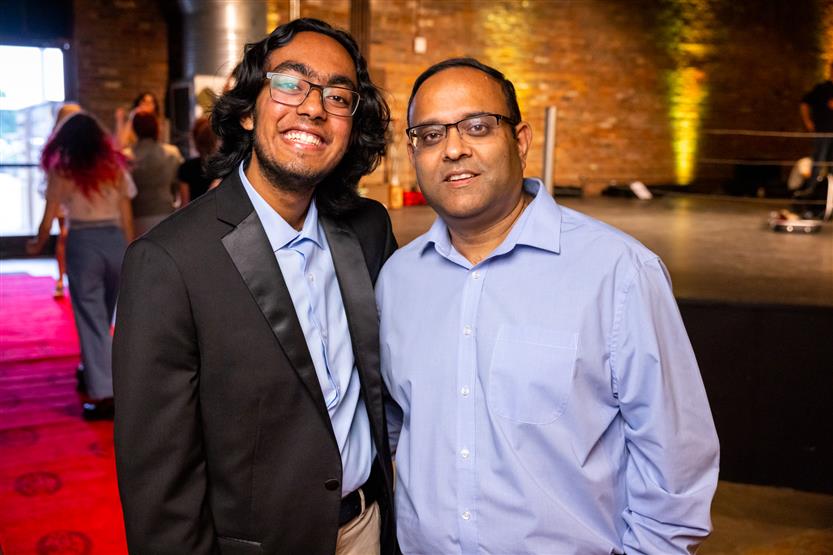 A young man wearing a black blazer and a man wearing a blue button up smile